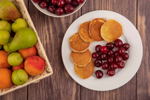 Bovenaanzicht van pannenkoeken met kersen in plaat en mand met fruit als peren-abrikozenpruim op houten achtergrond