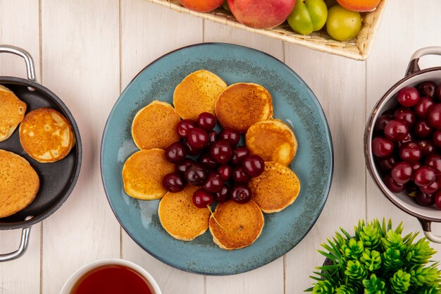 Bovenaanzicht van pannenkoeken met kersen in plaat en pan pannenkoeken met kom met kersen en fruit met thee op houten achtergrond