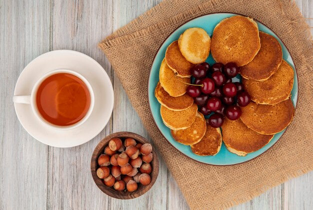 Bovenaanzicht van pannenkoeken met kersen in plaat op zak en kopje thee met kom met noten op houten achtergrond