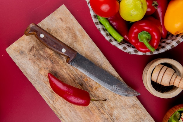 Bovenaanzicht van peper en mes op snijplank met groenten in mand en knoflook crusher op bordo oppervlak