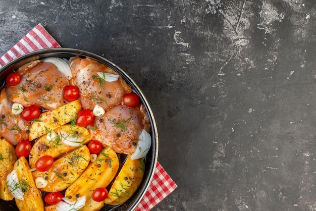 Bovenaanzicht van pittige rauwe kippenmaaltijd met aardappelen, groenten in pan op rode gestripte handdoek aan de rechterkant op grijze achtergrond