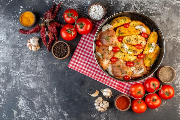 Bovenaanzicht van pittige rauwe kippenmaaltijd met aardappelen groenten in pan op rode gestripte handdoek en gedroogde pepers knoflook tomaten gele gember op grijze achtergrond