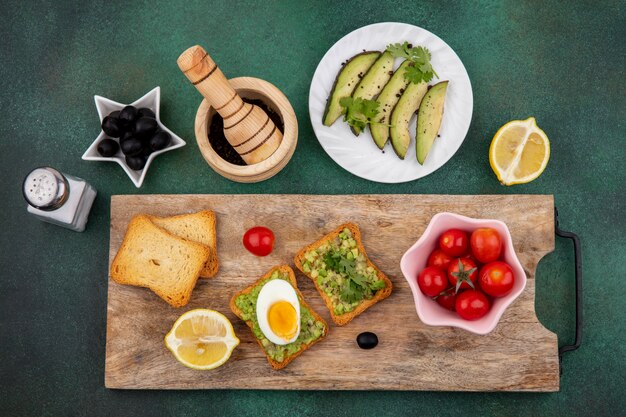 Bovenaanzicht van plakjes avocado op witte plaat met een geroosterde sneetjes brood met avocadopulp en ei op houten keukenbord met tomaten op roze kom op gre