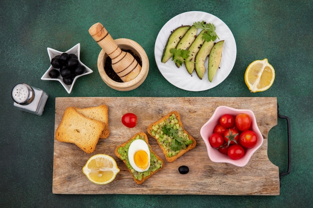 Bovenaanzicht van plakjes avocado op witte plaat met een geroosterde sneetjes brood met avocadopulp en ei op houten keukenbord met tomaten op roze kom op gre