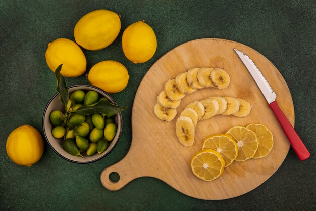 Bovenaanzicht van plakjes vers fruit zoals bananen en citroenen op een houten keukenbord met mes met kinkans op een kom met citroenen geïsoleerd op een groene achtergrond