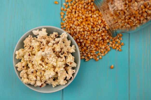 Bovenaanzicht van popcorns pitten vallen uit een glazen pot met lekkere popcorns op een kom op een blauwe houten tafel