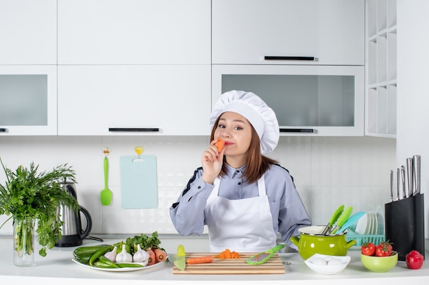 Bovenaanzicht van positieve chef-kok en verse groenten met kookgerei en proeverij van wortel in de witte keuken