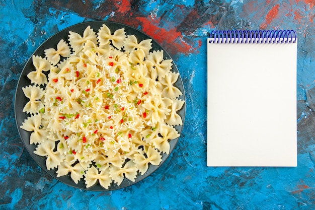 Bovenaanzicht van rauwe Italiaanse farfalle pasta met groenten en spiraalvormig notitieboekje op blauwe achtergrond