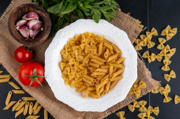 Bovenaanzicht van rauwe pasta in een bord met tomaten, knoflook en een bosje munt op een beige servet
