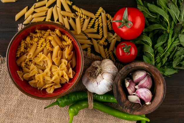 Bovenaanzicht van rauwe pasta in een kom met tomaten, knoflook, chili peper en een bosje munt op een beige servet