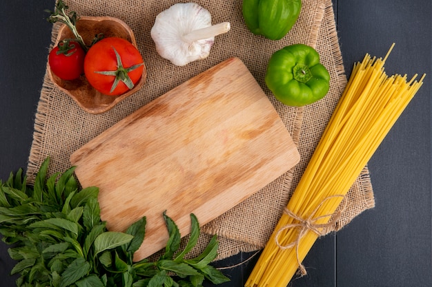 Bovenaanzicht van rauwe pasta met snijplank tomaten knoflook munt en paprika op een beige servet