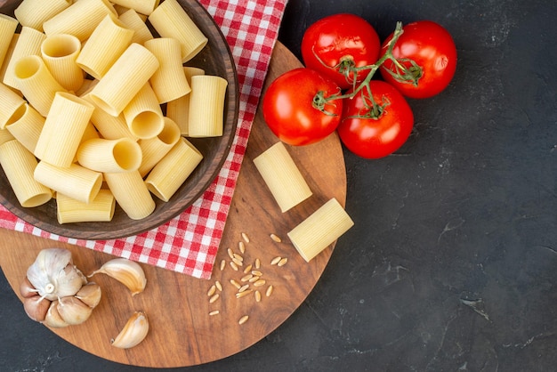 Bovenaanzicht van rauwe pasta's binnen en buiten een bruine kom op rode gestripte handdoek knoflook rijst op ronde houten plank tomaten op zwarte achtergrond