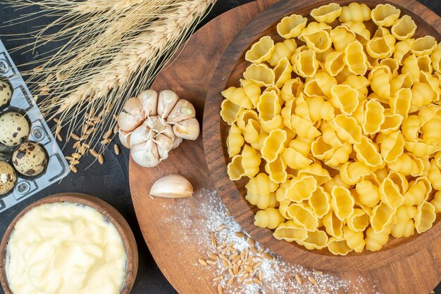 Bovenaanzicht van rauwe pasta's in een bruine kom knoflook rijst op houten plank meel eieren spikes op zwarte achtergrond