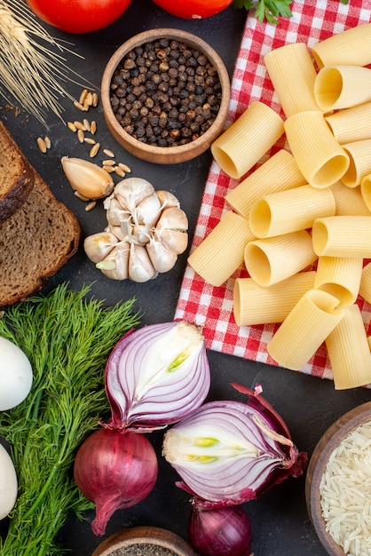 Bovenaanzicht van rauwe pasta's op rode gestripte handdoekrijst in een bruine kom, verse groenten, sneetjes brood op zwarte tafel