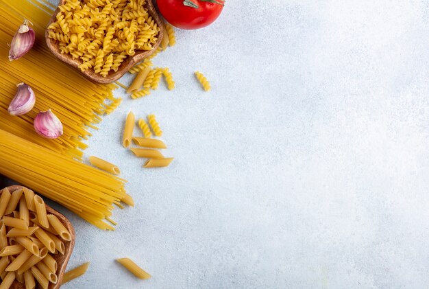 Bovenaanzicht van rauwe spaghetti met rauwe pasta in kommen met knoflook en tomaten op een grijze ondergrond