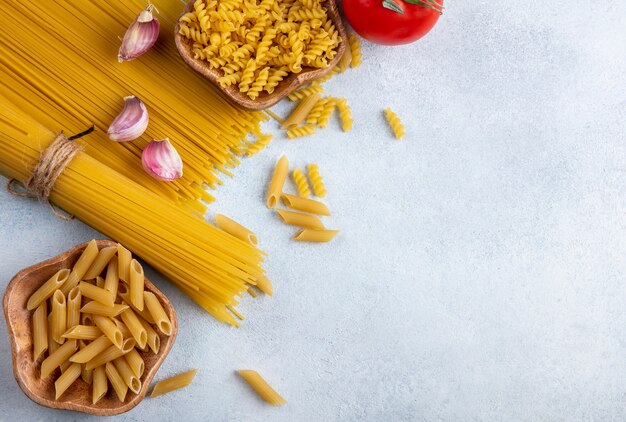 Bovenaanzicht van rauwe spaghetti met rauwe pasta in kommen met knoflook en tomaten op een grijze ondergrond