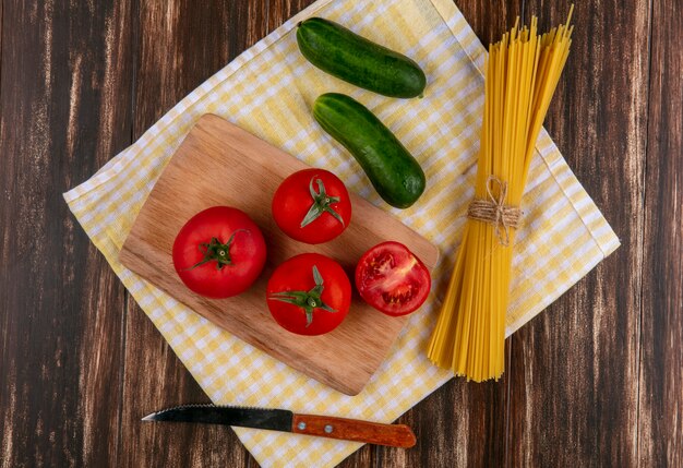 Bovenaanzicht van rauwe spaghetti met tomaten op een snijplank met een mes en komkommers op een gele geruite handdoek op een houten oppervlak