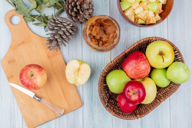 Bovenaanzicht van rode appel en mes op snijplank met mandje met appels potje appeljam kom appelblokjes dennenappels en bladeren op houten achtergrond