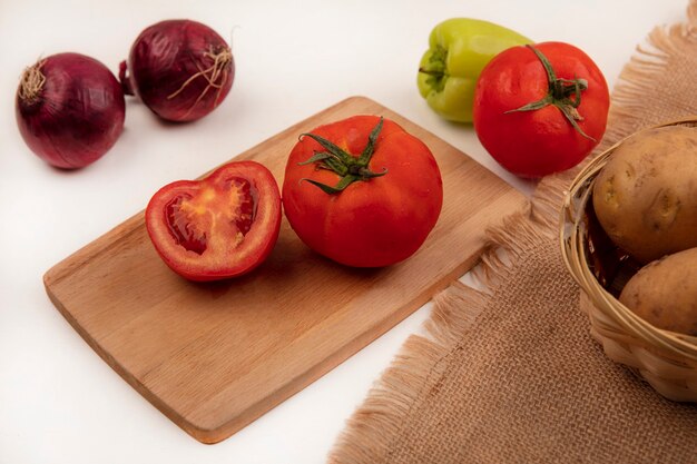 Bovenaanzicht van rode tomaten op een houten keukenbord met verse aardappelen op een emmer op een zakdoek met rode uien en groene peper geïsoleerd op een witte muur