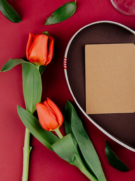 Bovenaanzicht van rode tulp bloemen met hartvormige geschenkdoos met een open briefkaart op rode achtergrond