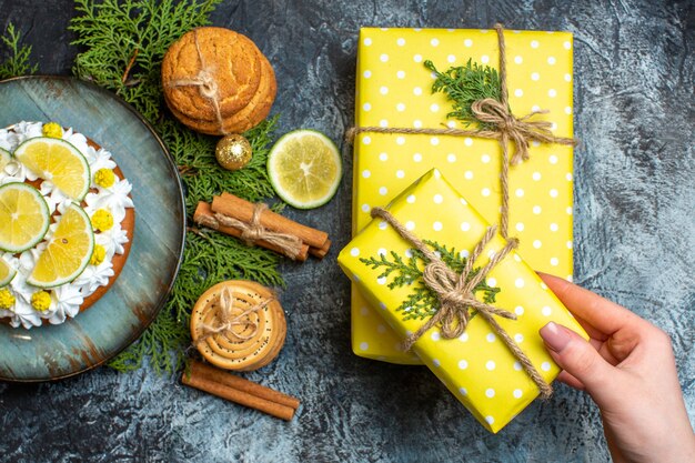 Bovenaanzicht van romige heerlijke cake en dennentakken, citroen, kaneel, limoenen, hand met een van de gele geschenkdozen op een donkere achtergrond