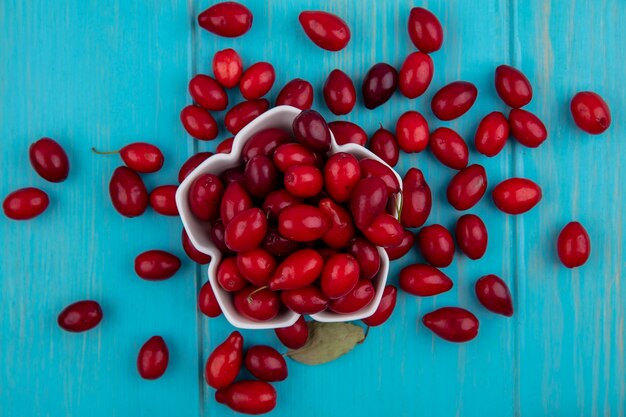 Bovenaanzicht van rood en vers cornel fruit op een witte kom op een blauwe houten achtergrond