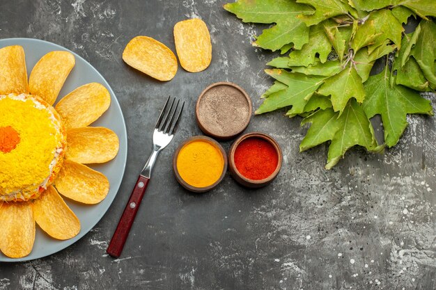 Bovenaanzicht van salade aan de linkerkant met kruiden, vork, chips en bladeren aan de zijkant op donkergrijze tafel