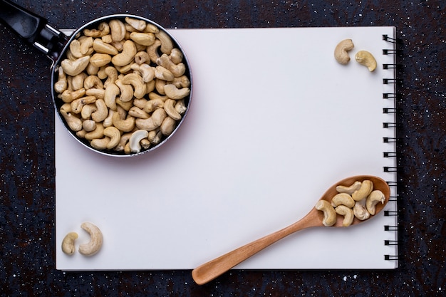 Bovenaanzicht van schetsboek en een pan met cashew en een houten lepel met noten op zwarte achtergrond