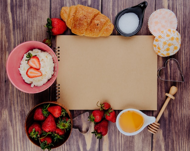 Bovenaanzicht van schetsboek en verse rijpe aardbeien met honing, suiker, croissant, kwark en koekjessnijders op hout