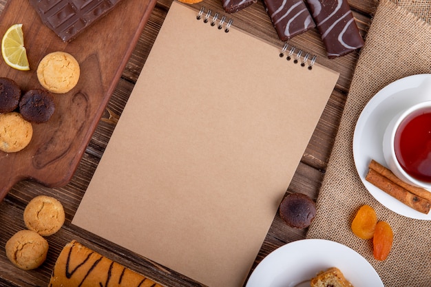 Bovenaanzicht van schetsboek verschillende zoete gebakjes cookies en een kopje thee met kaneelstokjes op houten achtergrond