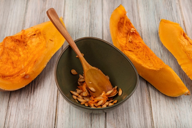 Bovenaanzicht van smakelijke oranje pompoenplakken met zaden op een kom met houten lepel op een grijze houten oppervlakte