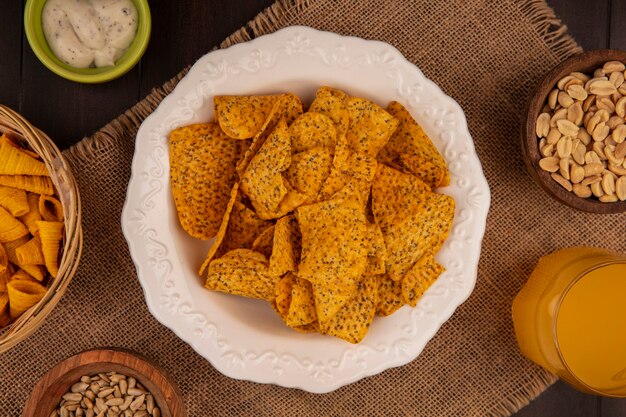 Bovenaanzicht van smakelijke pittige chips op een witte kom op een zakdoek met pijnboompitten op een houten kom met gepelde zonnebloempitten met een glas sinaasappelsap op een houten tafel