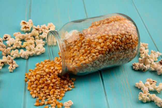 Bovenaanzicht van smakelijke popcorn-pitten die uit een glazen pot vallen met popcorns geïsoleerd op een blauwe houten tafel