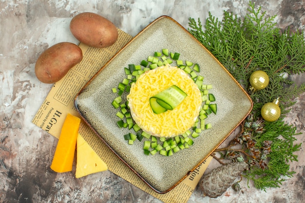 Bovenaanzicht van smakelijke salade geserveerd met gehakte komkommer op een oude krant en twee soorten kaas en wortelen aardappelen op gemengde kleur achtergrond