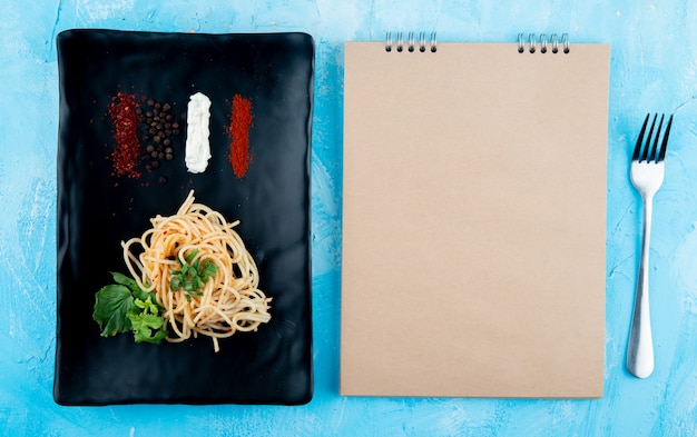 Bovenaanzicht van spaghetti pasta met basilicum en specerijen op een zwarte schotel en schetsboek op blauwe achtergrond