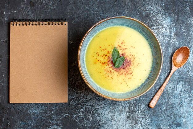 Bovenaanzicht van spiraalvormige notebook heerlijke soep geserveerd met peper en munt in een blauwe pot naast houten lepel op donkere achtergrond