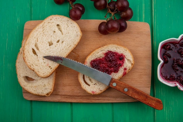 Bovenaanzicht van stokbrood segment besmeurd met frambozenjam met mes op snijplank en druivenmost met kom frambozenjam op groene achtergrond