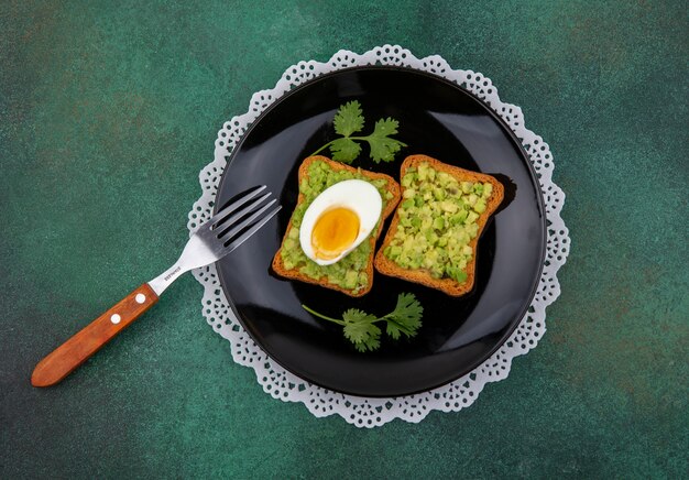Bovenaanzicht van toast sneetjes brood met avocado pulps en ei op een kom met vork op groene ondergrond
