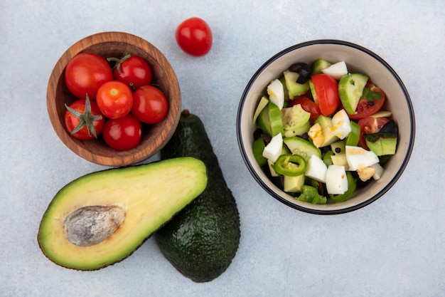 Bovenaanzicht van tomaten in een houten kom met avocado in de buurt van plantaardige salade in een kom op witte ondergrond