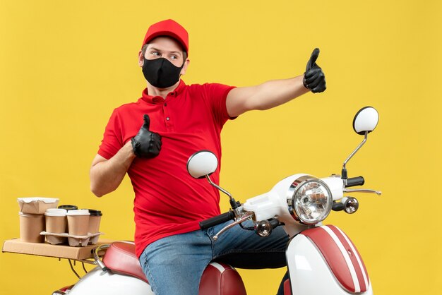 Bovenaanzicht van trotse zelfverzekerde jonge volwassene die rode blouse en muts handschoenen in medisch masker draagt ?? die bestelling op scooter op gele achtergrond levert