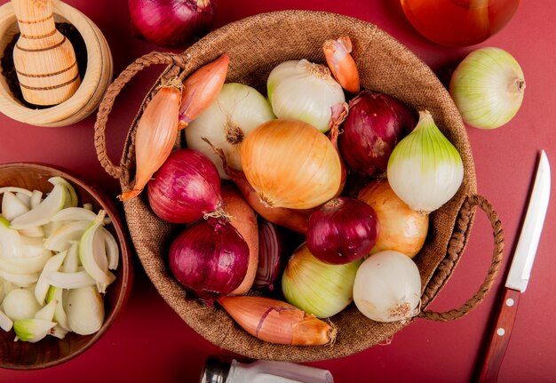 Bovenaanzicht van uien in mand met zwarte peper, gesneden ui in kom, zout, boter en mes rond op rood oppervlak