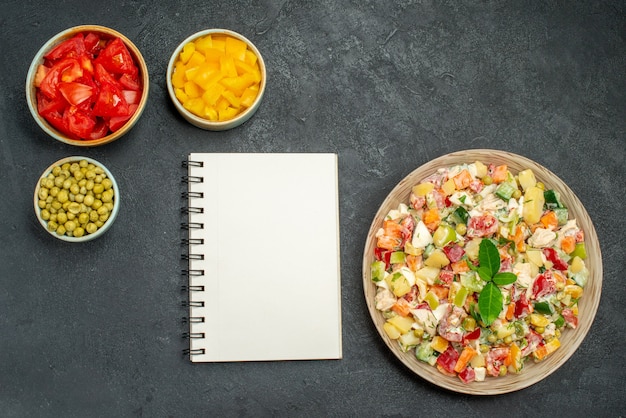 Bovenaanzicht van vegetarische salade met kommen met groenten en blocnote aan kant op donkergrijze achtergrond