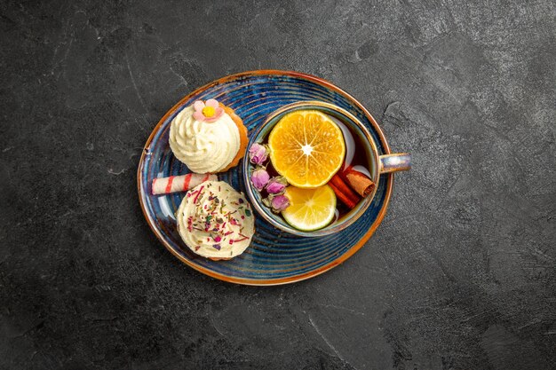 Bovenaanzicht van ver een kopje thee met citroen twee cupcakes met room en een kopje kruidenthee met citroen op de blauwe schotel op de donkere tafel