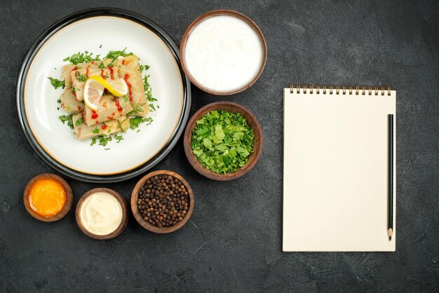 Bovenaanzicht van ver eten op tafel kommen van zure room kruiden zwarte peper en gele saus en gevulde kool met kruiden citroen en saus op witte plaat naast wit notitieboekje en potlood op zwarte tafel