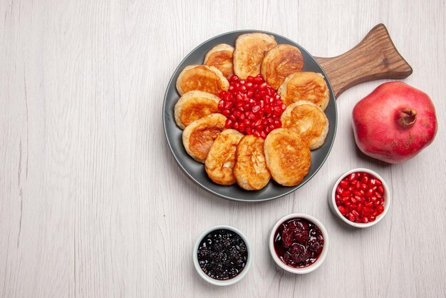 Bovenaanzicht van ver lekker gerecht granaatappel kommen jam en bord smakelijke pannenkoeken en granaatappel op de houten snijplank op tafel