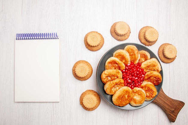 Gratis foto bovenaanzicht van veraf bord op het bord koekjes en bord met smakelijke pannenkoeken en granaatappel op de houten snijplank naast het witte notitieboekje op tafel