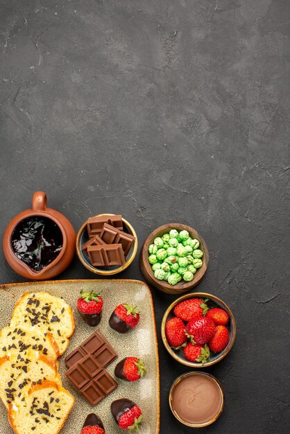 Bovenaanzicht van veraf chocolade aardbeien cake chocolade aardbeien groene snoepjes en chocolade crème in kommen smakelijke cake en aardbeien op de donkere tafel