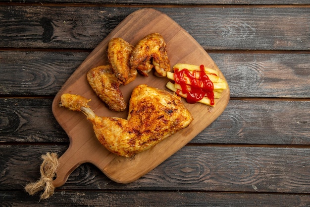 Bovenaanzicht van veraf kippenvleugels en been met frietjes en ketchup op de houten snijplank op de donkere tafel