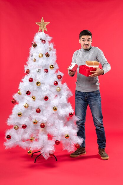 Bovenaanzicht van verrast knappe volwassene in een grijze blouse staande in de buurt van de versierde witte kerstboom