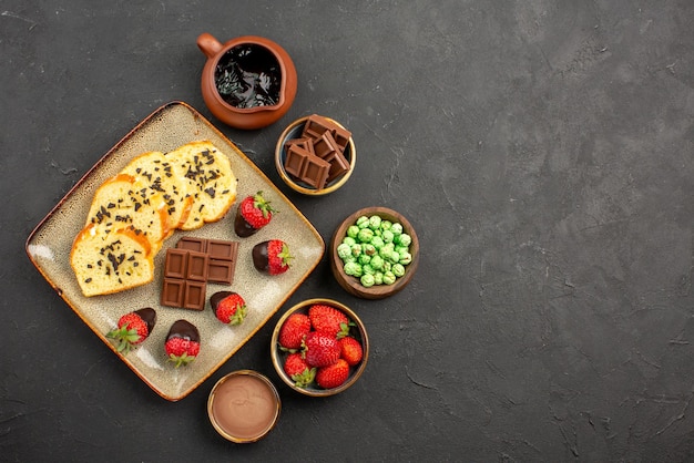 Bovenaanzicht van verre cake en aardbeien kommen met chocolade aardbeien, groene snoepjes en chocoladeroom naast het bord cake met met chocolade omhulde aardbeien